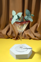 Photo of Beautiful martini glass with eucalyptus leaves on yellow surface against brown curtain