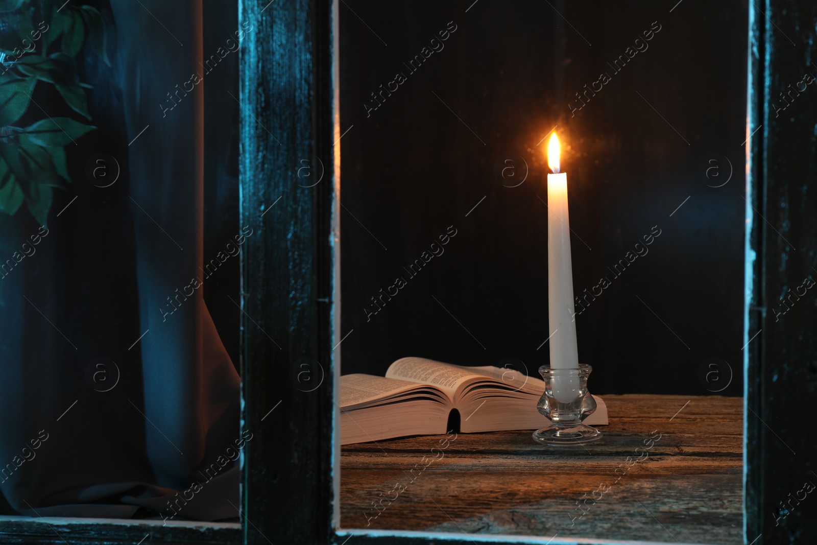 Photo of Burning candle and Bible on wooden table at night, view through window