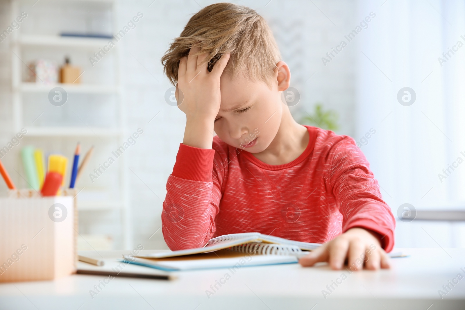 Photo of Little boy suffering from headache while doing homework at table indoors