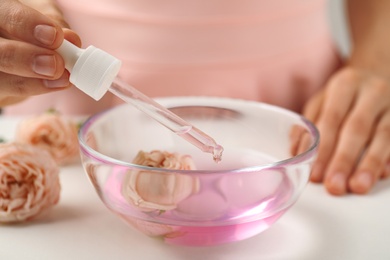 Photo of Young woman dripping rose essential oil into glass bowl at table, closeup