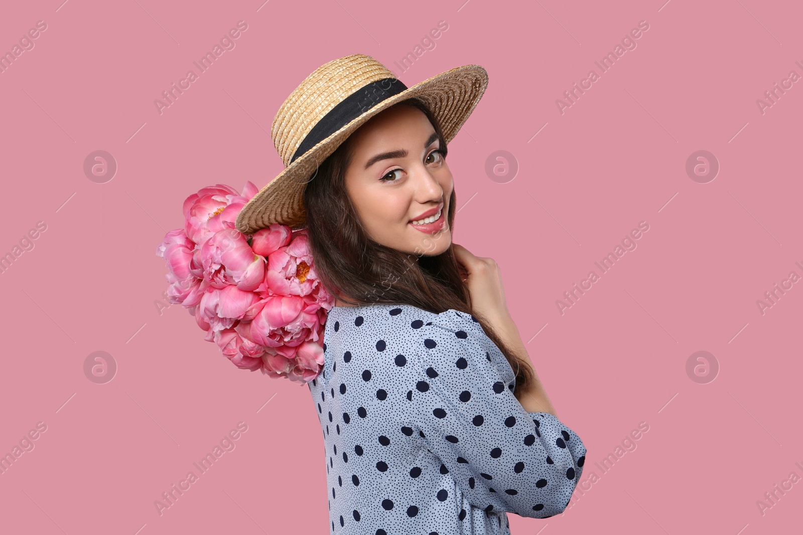 Photo of Beautiful young woman in straw hat with bouquet of peonies on pink background