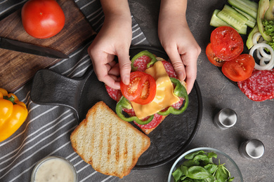 Photo of Woman adding tomato to sandwich at grey table, top view