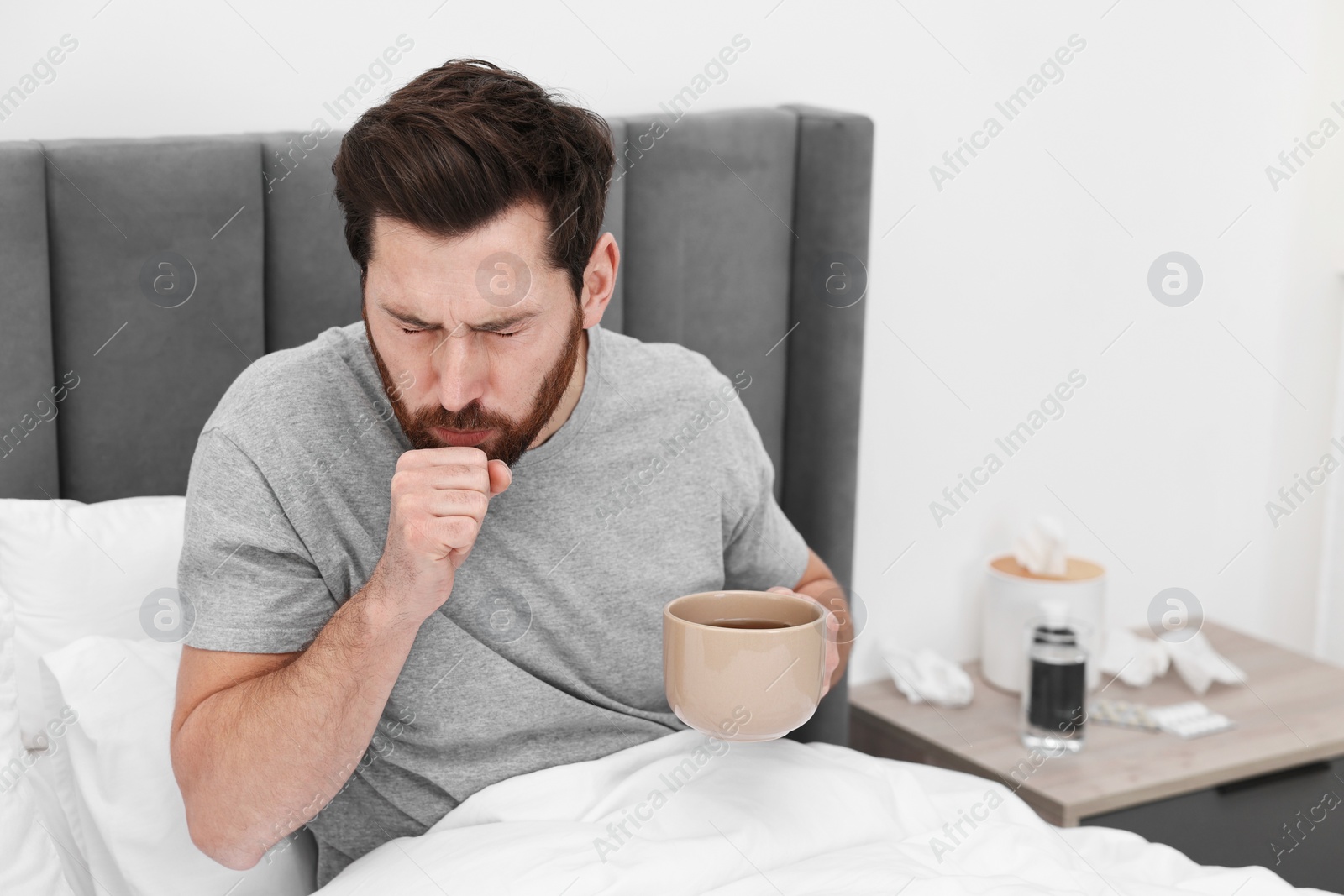 Photo of Sick man with cup of hot drink coughing on bed at home. Cold symptoms