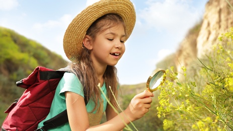 Cute little girl exploring plant with magnifying glass outdoors on sunny day