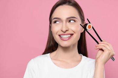 Beautiful young woman holding sushi roll with chopsticks on pink background