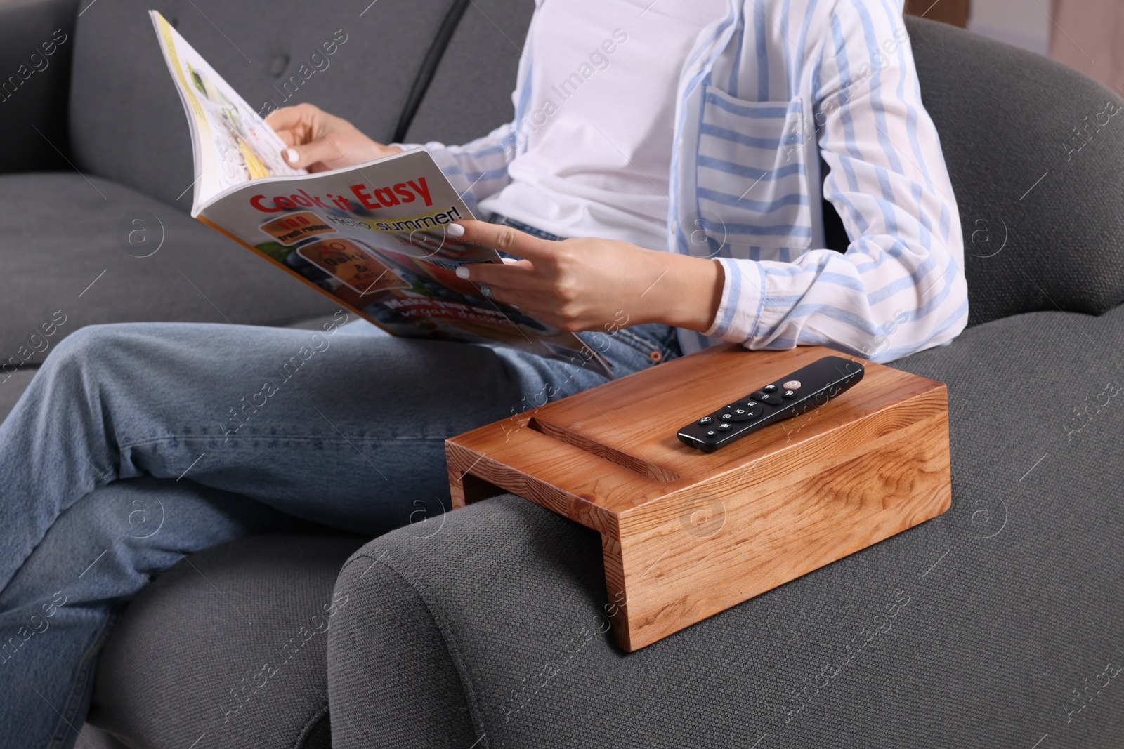 Photo of Remote control on sofa armrest wooden table. Woman reading magazine at home, closeup