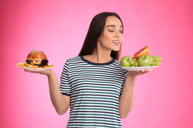 Photo of Woman choosing between fruits and burger with French fries on pink background
