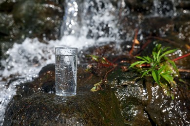 Photo of Wet glass of water on rocks near flowing stream outdoors, space for text