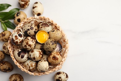 Wicker bowl with whole and cracked quail eggs on white marble table, flat lay. Space for text