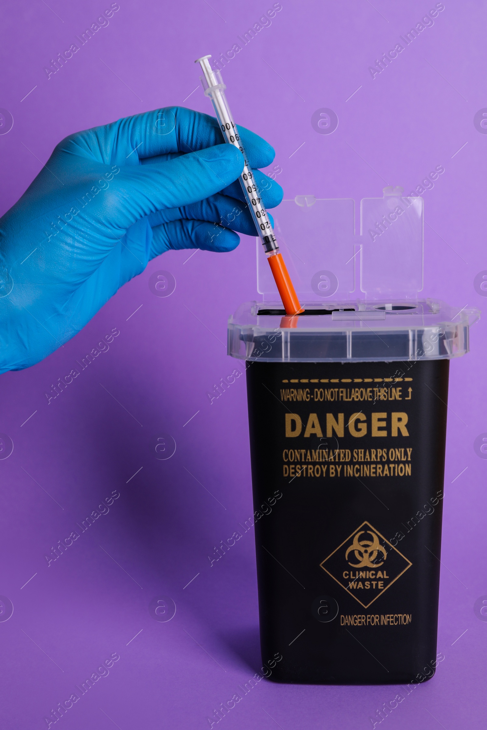 Photo of Doctor throwing used syringe into sharps container on violet background, closeup