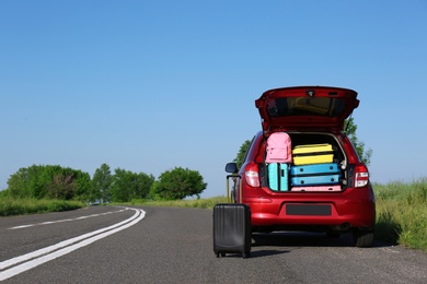 Photo of Suitcase near family car with open trunk full of luggage on highway. Space for text