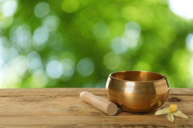 Image of Golden singing bowl, mallet and flower on wooden table against outdoors, space for text