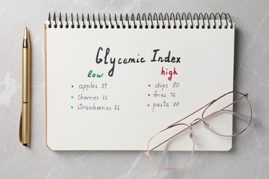 Photo of List with products of low and high glycemic index in notebook, pen and glasses on light grey marble table, top view