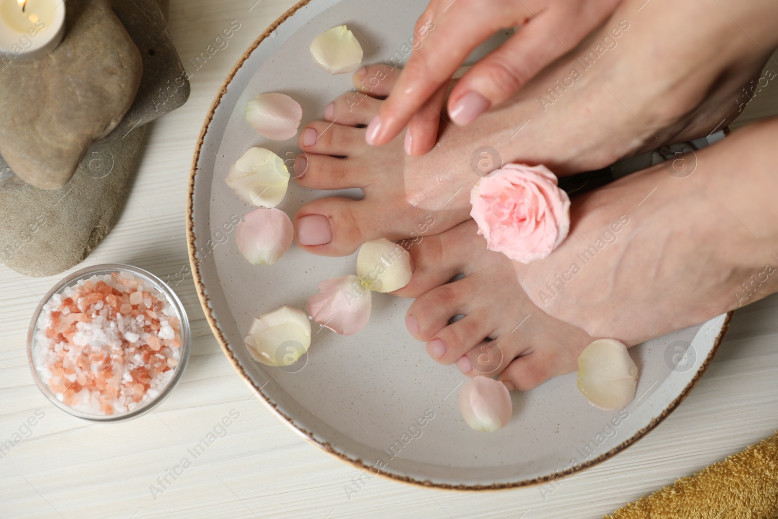 Photo of Woman soaking her feet in bowl with water, flower and petals on white wooden floor, top view. Pedicure procedure