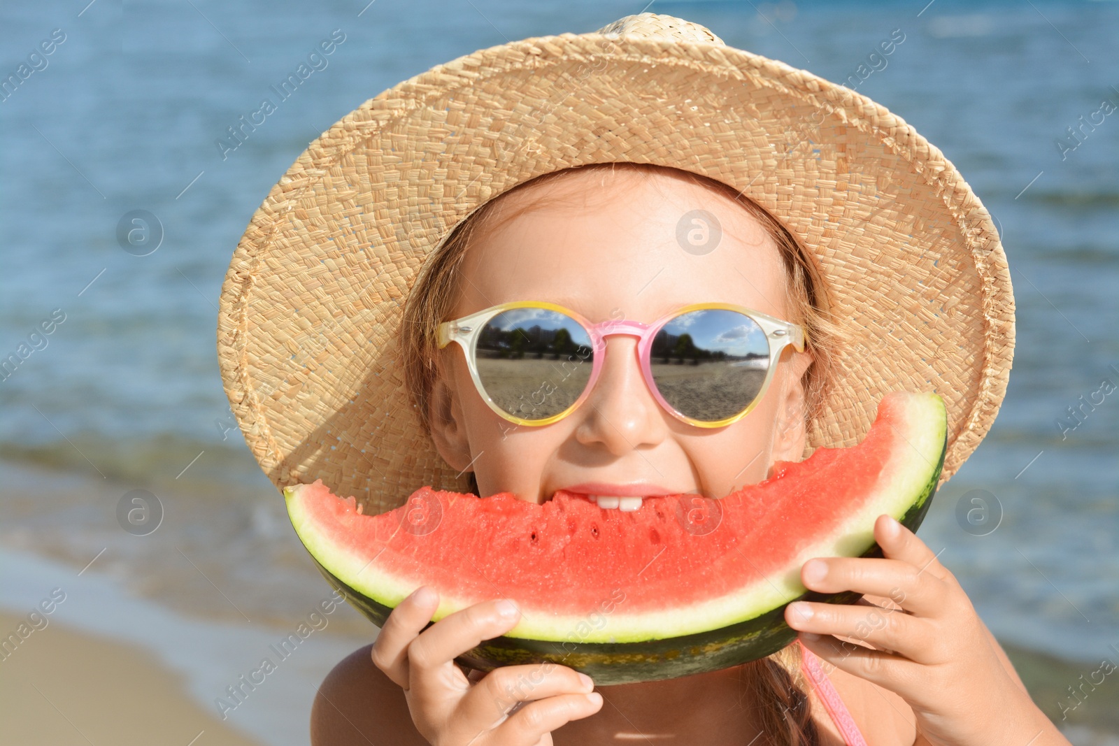 Photo of Cute little girl in straw hat and sunglasses eating juicy watermelon on beach outdoors