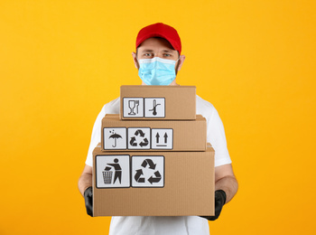 Photo of Courier in mask holding cardboard boxes with different packaging symbols on yellow background. Parcel delivery