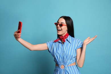 Photo of Fashionable young woman in stylish outfit with bandana taking selfie on light blue background
