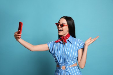 Fashionable young woman in stylish outfit with bandana taking selfie on light blue background