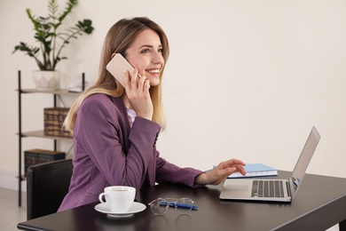 Photo of Young businesswoman talking on phone while using laptop at table in office