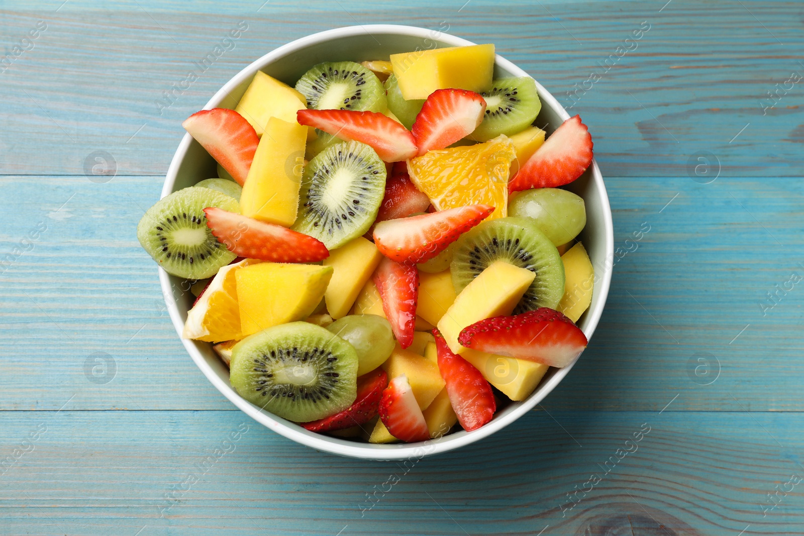 Photo of Tasty fruit salad in bowl on light blue wooden table, top view