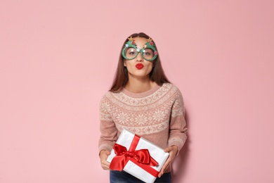 Young woman in warm sweater with Christmas gift and party glasses on color background