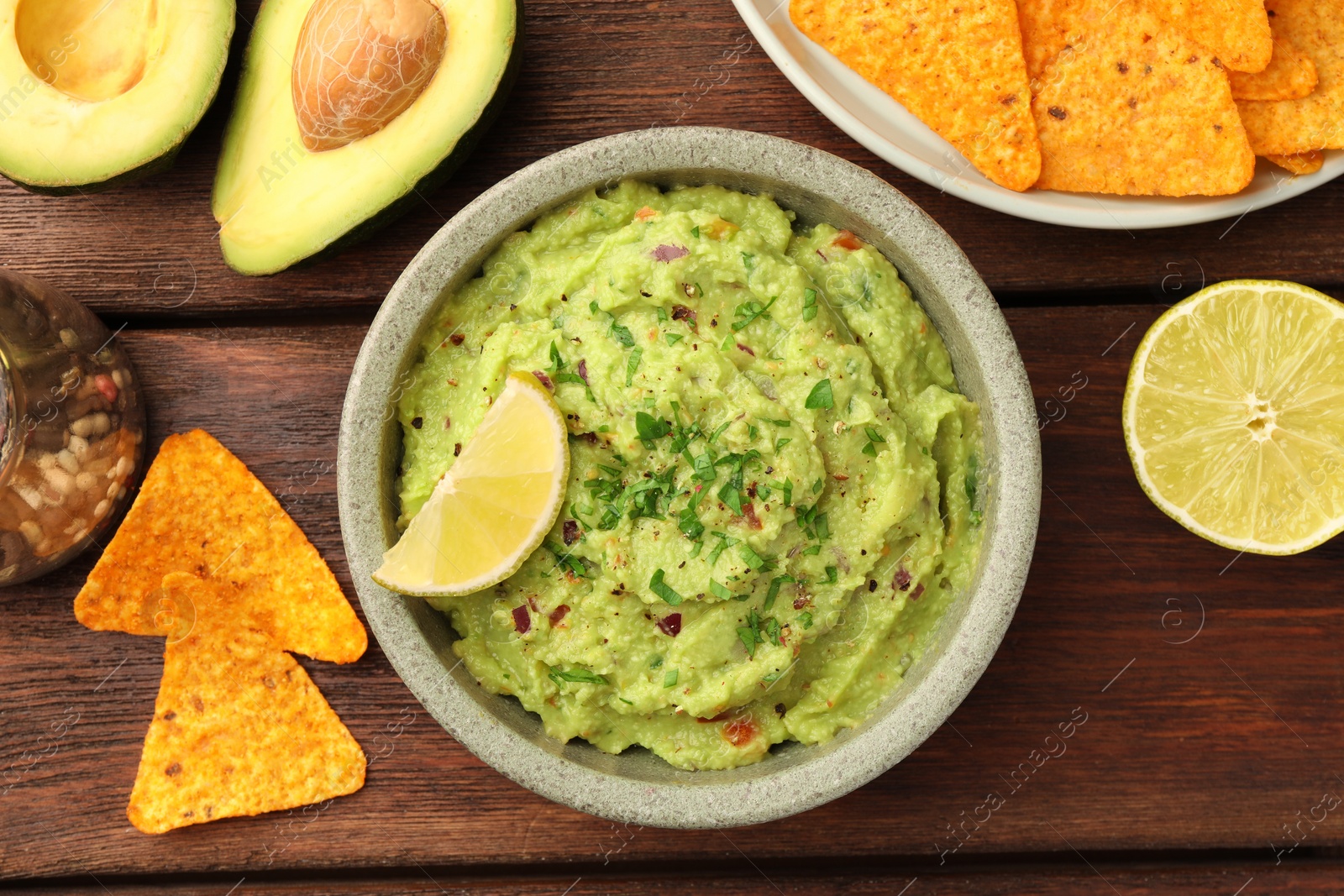Photo of Bowl of delicious guacamole, lime and nachos chips on wooden table, flat lay