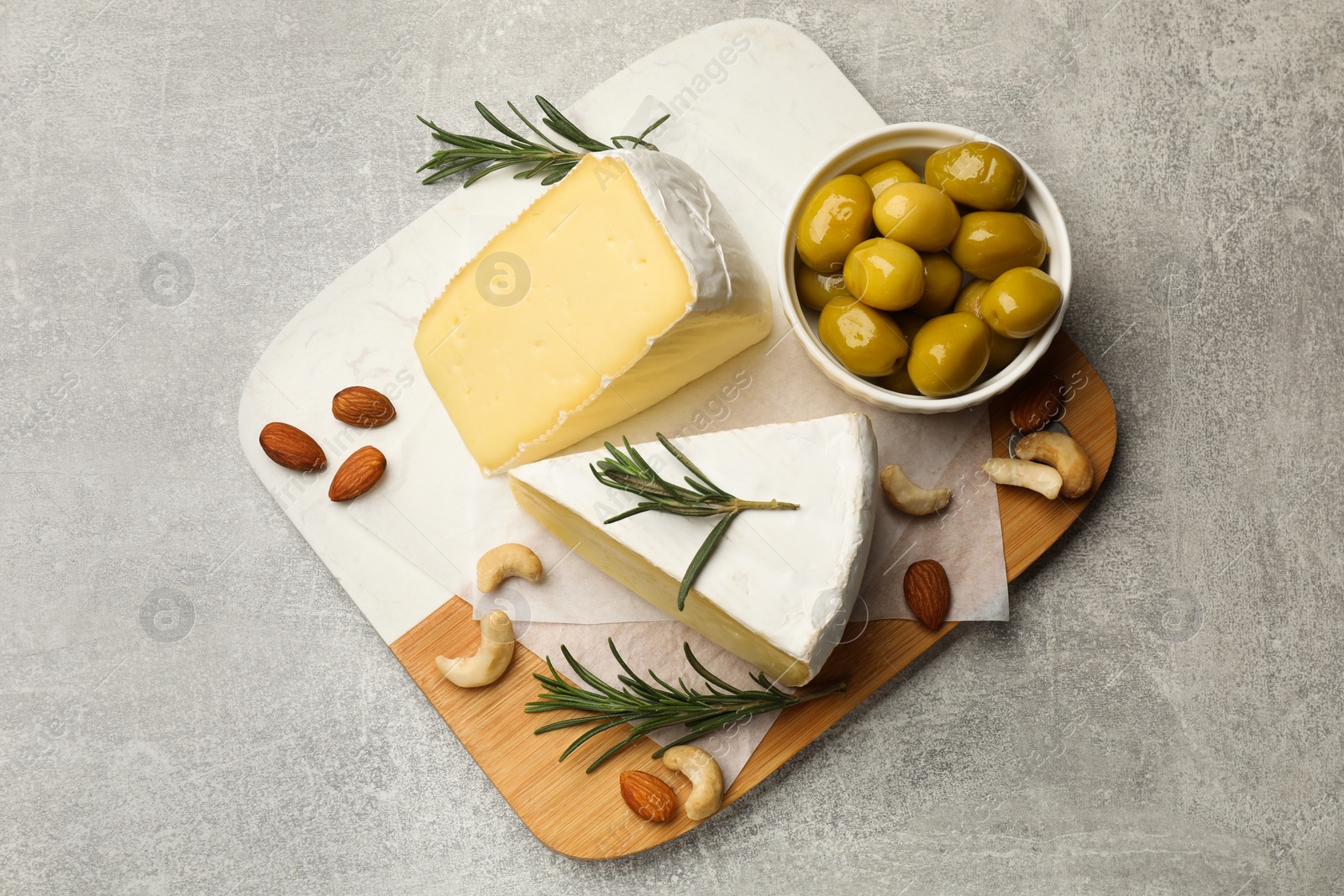 Photo of Pieces of tasty camembert cheese, rosemary, nuts and olives on grey textured table, top view