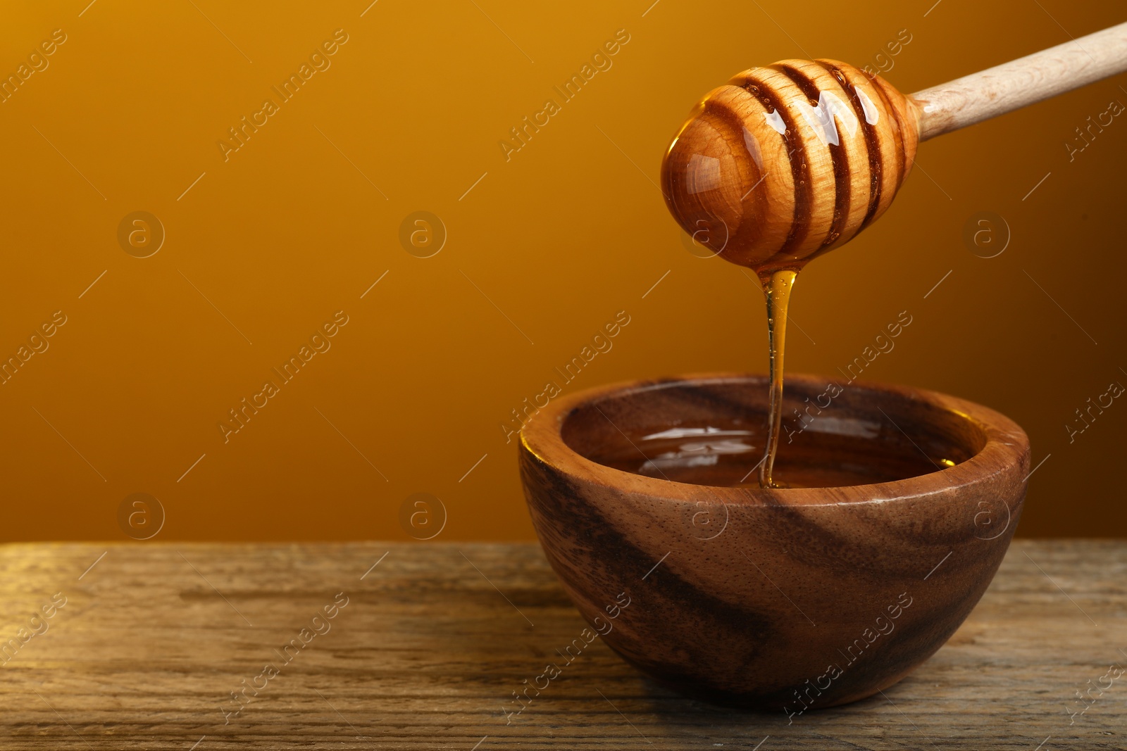 Photo of Pouring honey from dipper into bowl at wooden table against golden background, space for text