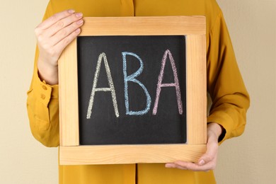 Photo of Woman holding small chalkboard with abbreviation ABA (Applied behavior analysis) on beige background, closeup
