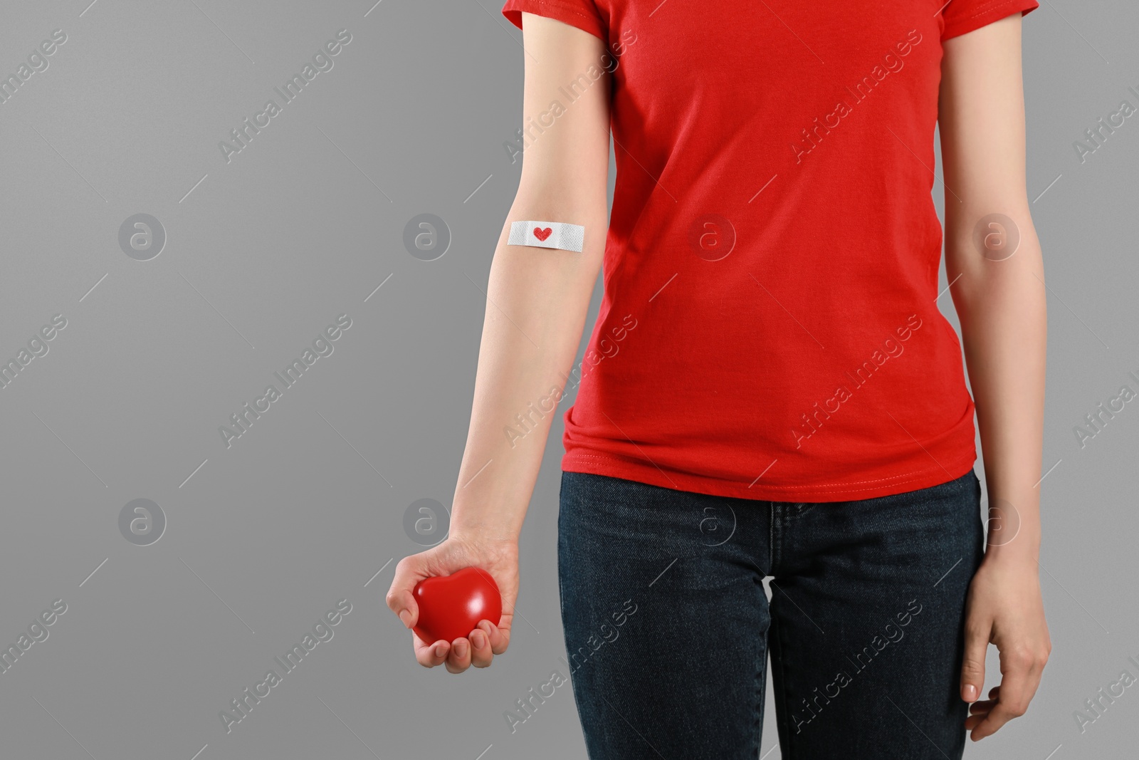 Photo of Blood donation concept. Woman with adhesive plaster on arm holding red heart against grey background, closeup. Space for text