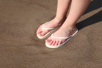 Photo of Woman in stylish white flip flops standing on wet sand, closeup