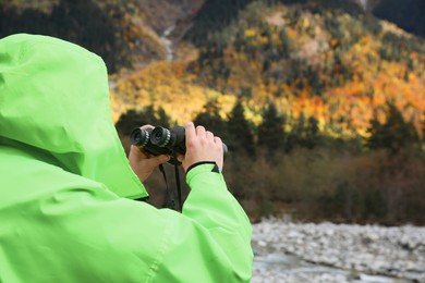 Photo of Woman holding binoculars near river in beautiful mountains, closeup. Space for text