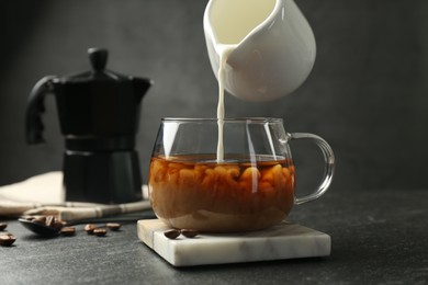 Pouring milk from pitcher into glass cup with coffee at dark textured table, closeup