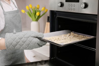 Photo of Woman putting baking sheet with homemade breadsticks into oven in kitchen, closeup. Cooking traditional grissini
