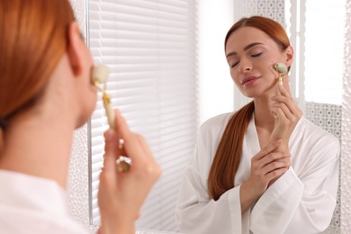 Photo of Young woman massaging her face with jade roller near mirror in bathroom