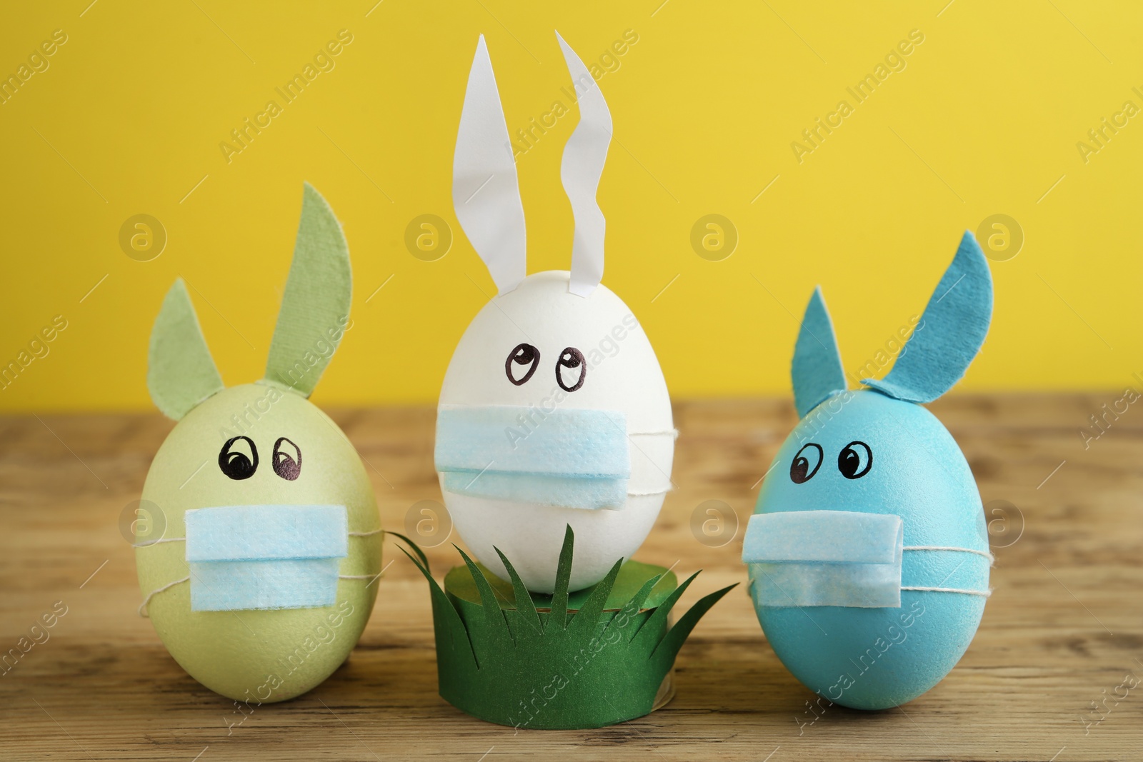 Photo of Dyed eggs with bunny ears in protective masks on wooden table against yellow background. Easter holiday during COVID-19 quarantine