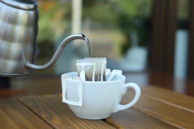 Photo of Pouring hot water into cup with drip coffee bag from kettle on wooden table, closeup