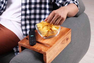 Photo of Man eating nacho chips on sofa with wooden armrest table at home, closeup
