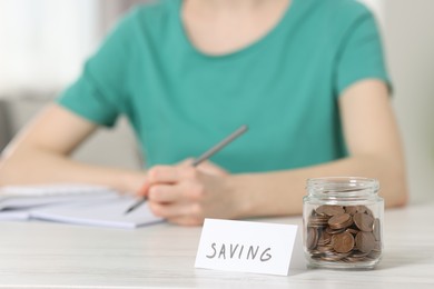 Photo of Financial savings. Woman making notes at white wooden table indoors, focus on glass jar with coins