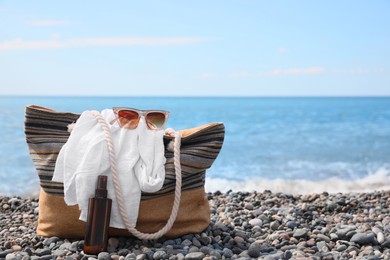 Beautiful bag with sunglasses, sunscreen and white shirt near sea on pebble beach. Space for text