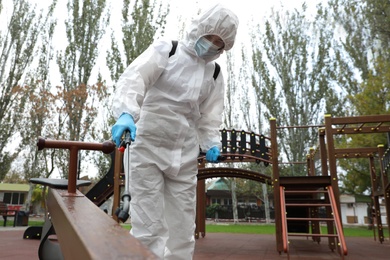 Photo of Woman wearing chemical protective suit with disinfectant sprayer on playground. Preventive measure during coronavirus pandemic