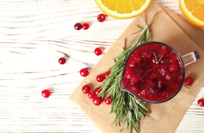 Photo of Tasty cranberry sauce in pitcher with rosemary on white wooden background, flat lay. Space for text