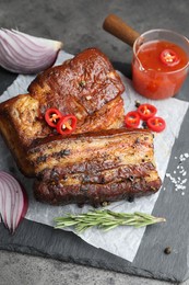 Photo of Pieces of baked pork belly served with sauce, rosemary and chili pepper on grey table