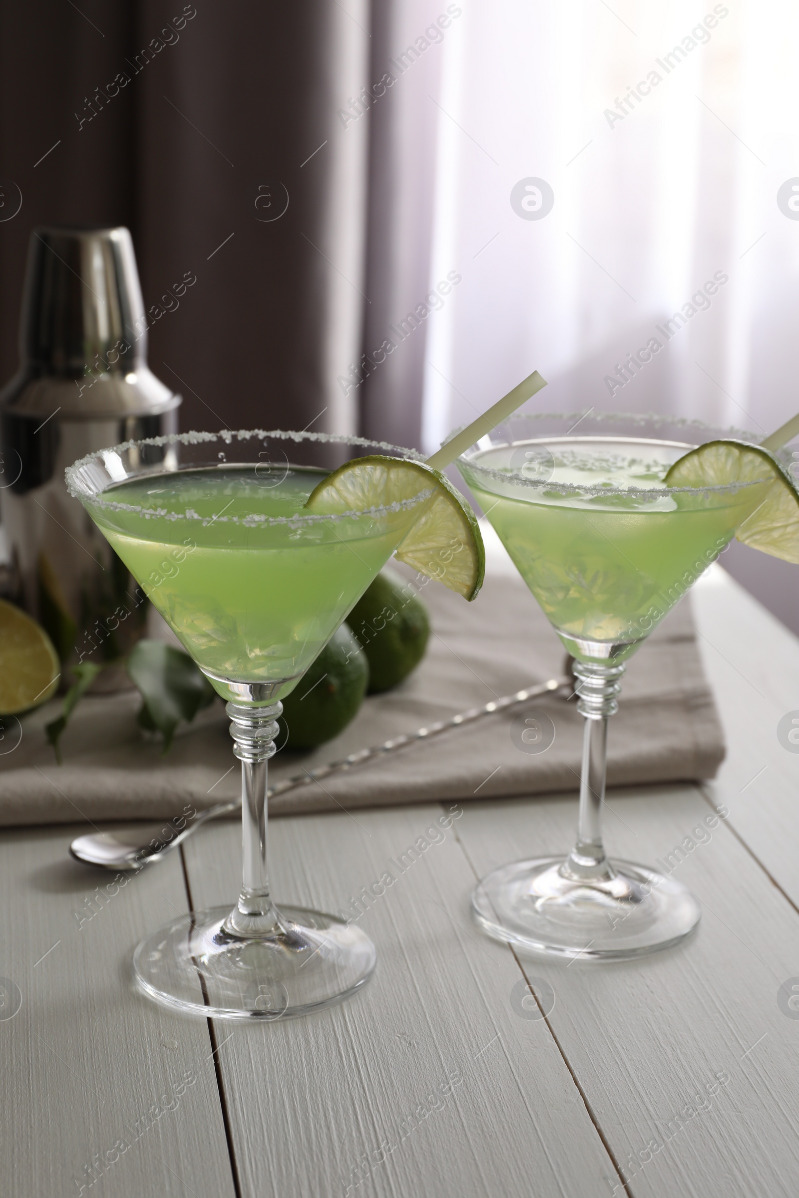 Photo of Delicious Margarita cocktail in glasses, limes, bar spoon and shaker on white wooden table