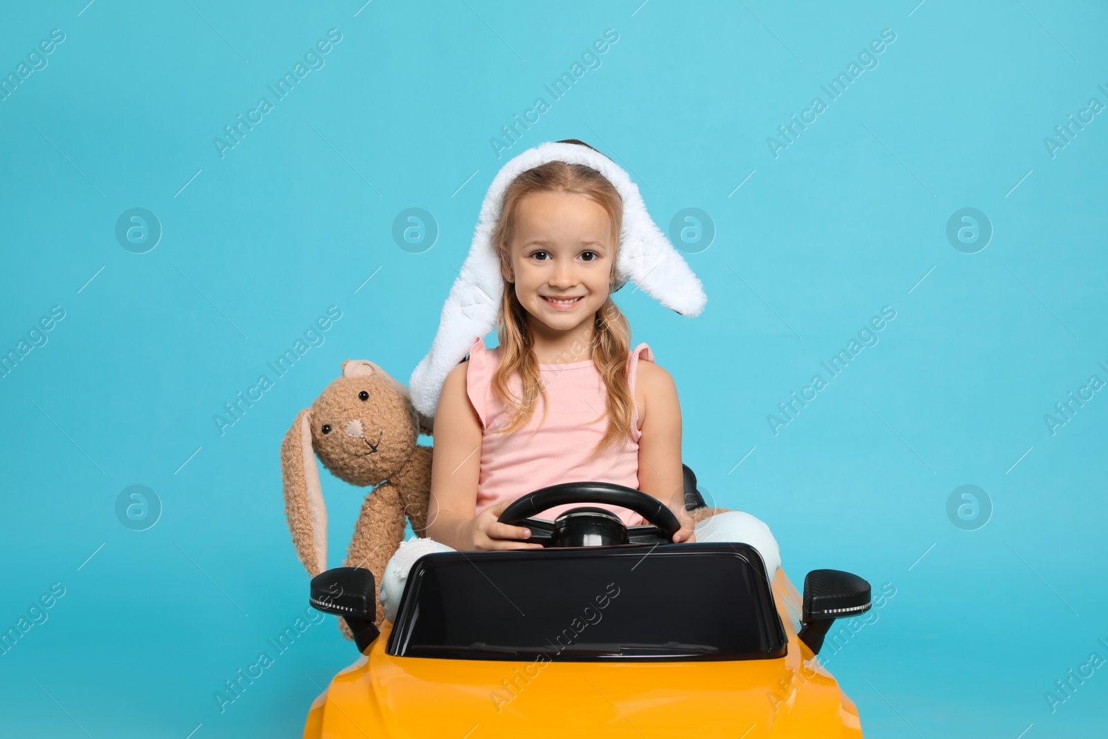 Photo of Cute little girl with toy bunny driving children's car on light blue background