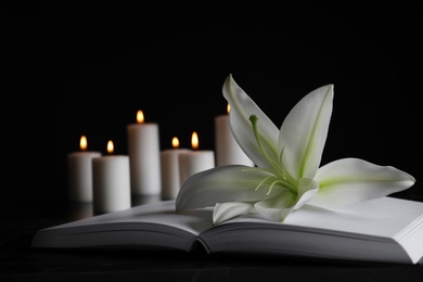 Photo of White lily, book and blurred burning candles on table in darkness, closeup with space for text. Funeral symbol