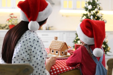 Photo of Mother and daughter decorating gingerbread house at table indoors, back view