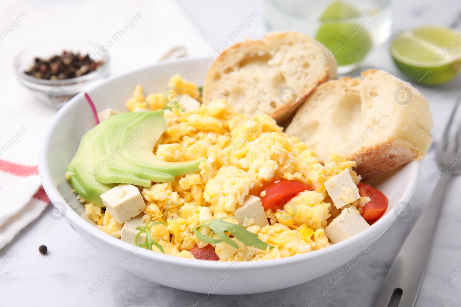 Photo of Bowl with delicious scrambled eggs, tofu, avocado and slices of baguette on white table, closeup