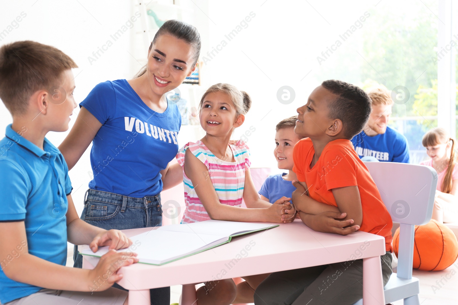 Photo of Young volunteer reading book with children at table indoors