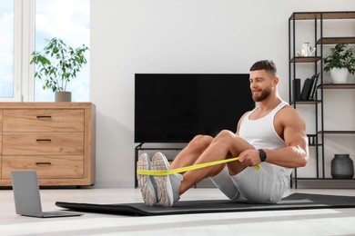 Muscular man doing exercise with elastic resistance band near laptop on mat at home